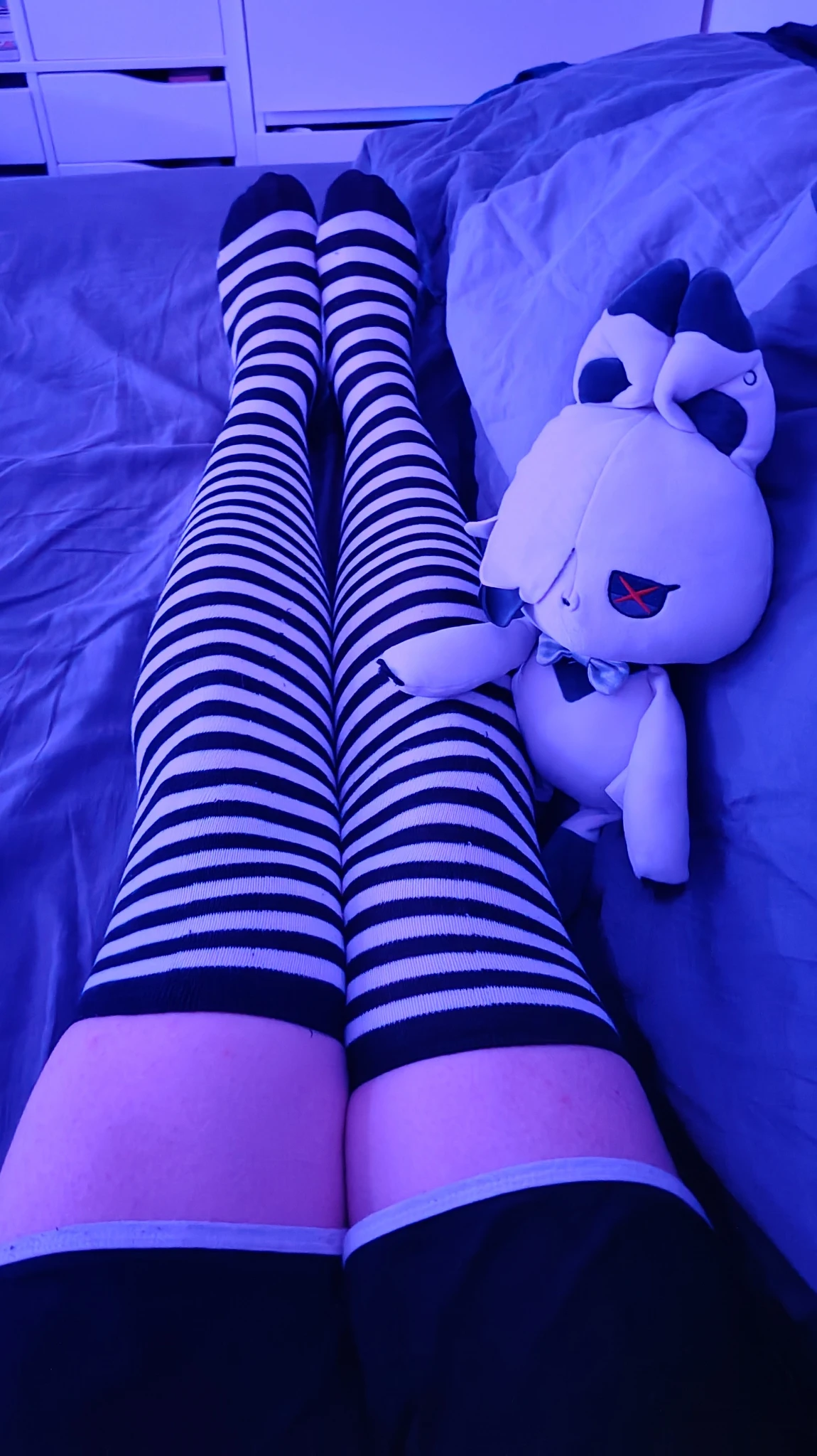 A first-person point-of-view shot looking down at legs wearing thigh-high, black-and-white horizontally striped socks under cool blue lighting. The person is lying on a messy bed, and a plush toy resembling Arlecchino from Genshin Impact as a bunny sits next to their right leg.