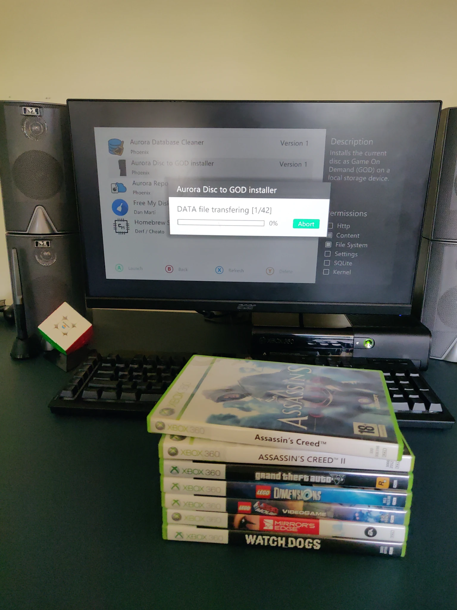 a picture of my desk with 7 Xbox 360 games stacked on top of each other in the following order from the top: Assassin's Creed, Assassin's Creed 2, Grand Theft Auto 5, LEGO Dimensions, The LEGO Movie Videogame, Mirror's Edge & Watch_Dogs. in the background on my monitor I am currently dumping the games to my HDD one by one using an Aurora Disc to GOD script.