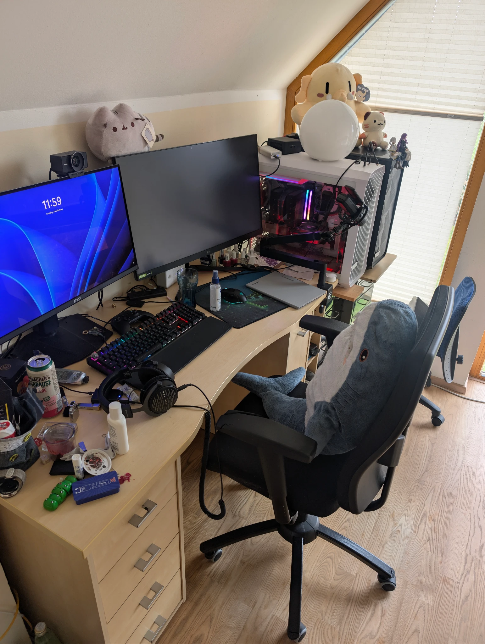 A cluttered gaming setup on a light wood desk in a room with sloped ceilings. Two monitors sit on the desk; the left one displays a blue Windows 11 lock screen showing "11:59." A large IKEA shark plush sits in a black gaming chair as if playing. The desk is crowded with a mechanical keyboard, a mouse, headphones. To the right, a white PC case with glowing internal RGB lights is topped with plush toys, including a large Pusheen and a round white lamp. A window with bright blinds is in the background.