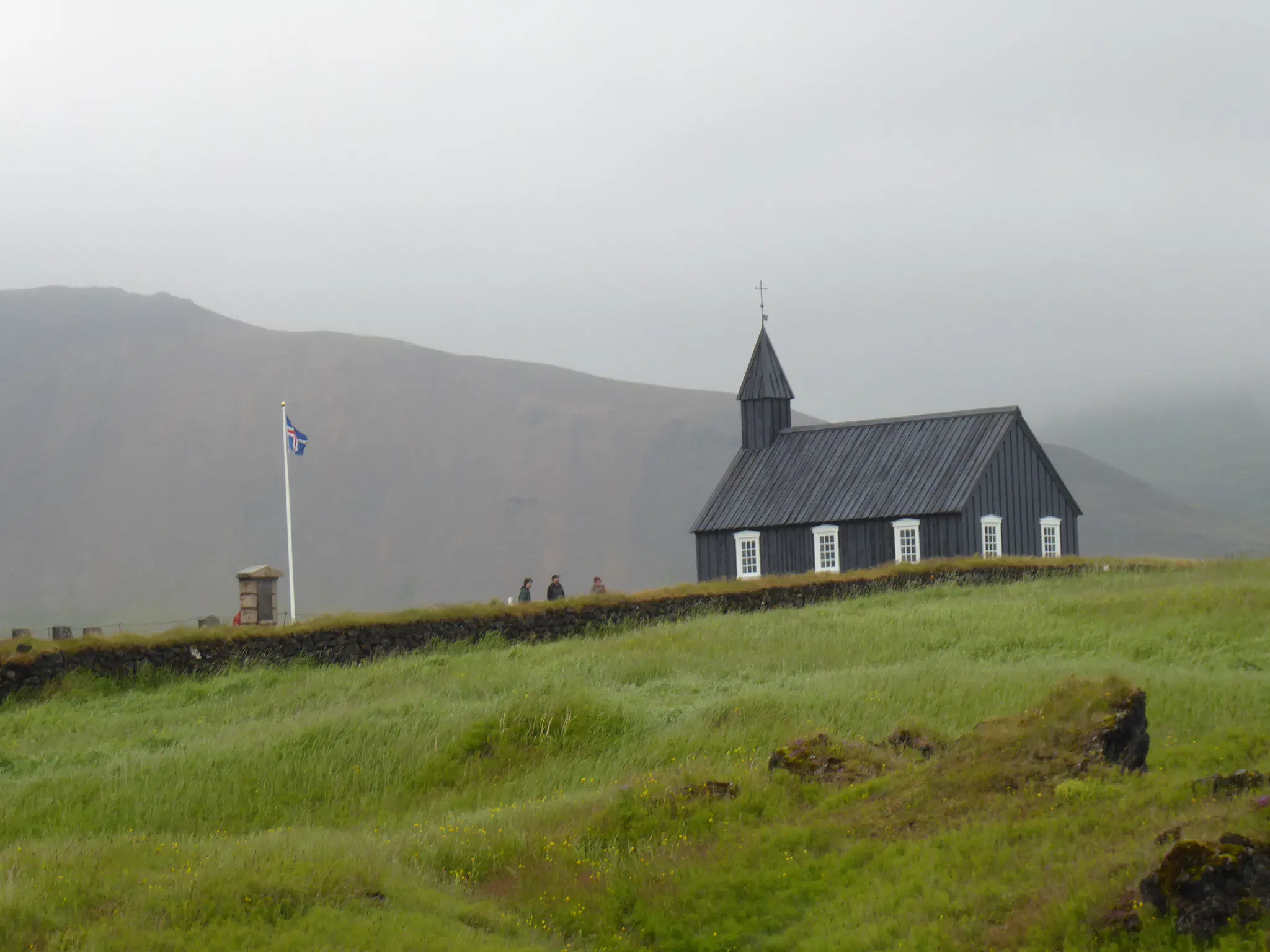 A small black wooden church in a green field. Misty hills in the background. A white flagpole with the Icelandic flag to the left.